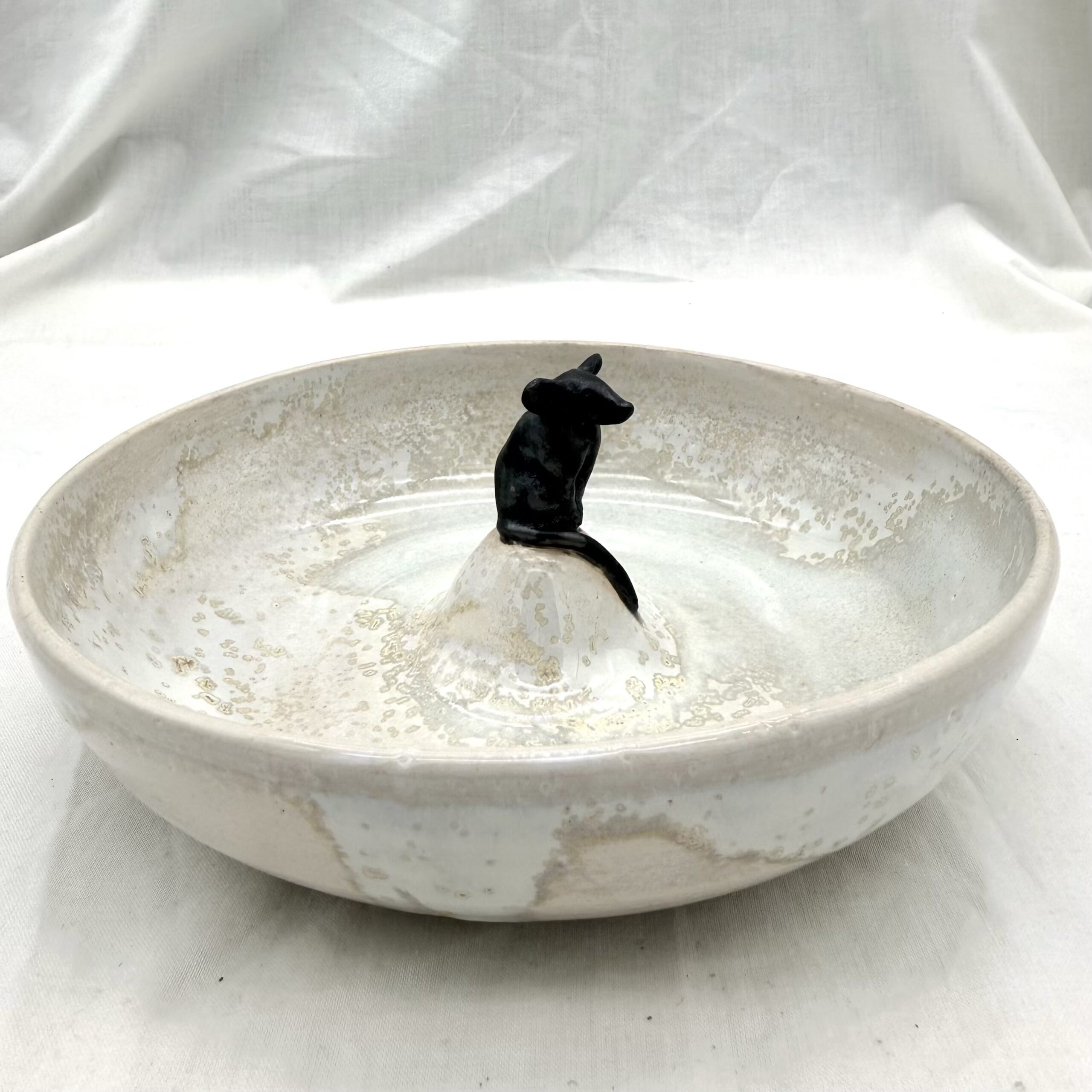 Bol, sur une île, petite souris Ce bol surmonté d’une souris noire égaiera votre intérieur et vos moments de convivialité. A usage multiple, pour apéritif ou collation, au aussi comme fontaine à eau pour les chats ou les oiseaux… Fabrique en grès blanc, au tour et émaillé en cristallisation sur fond blanc avec une finition brillante, la souris est modelée à la main et et émaillée en noir satiné. Chaque bol est une pièce unique et entièrement artisanale, il peut don y avoir d’une pièce à l’autre des variation de taille et de motif.